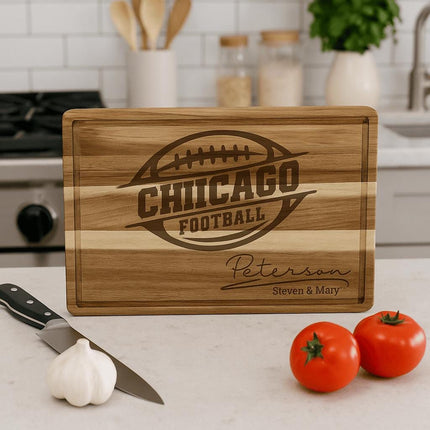 “Personalized Chicago football cutting board made from striped acacia wood, engraved with a football logo and the family name ‘Peterson – Steven & Mary.’ Displayed upright on a kitchen counter with garlic, tomatoes, a knife, and cooking utensils in the background, emphasizing its decorative and functional appeal.”