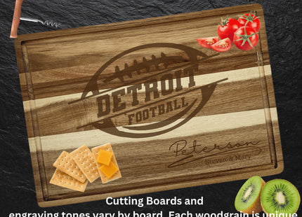 “Personalized Detroit football cutting board made from striped acacia wood, engraved with a football logo and the family name ‘Peterson – Steven & Mary.’ Displayed on a dark slate background with tomatoes, crackers with cheese, and sliced kiwi around it. Text at the bottom reads, ‘Cutting boards and engraving tones vary by board. Each woodgrain is unique.’”