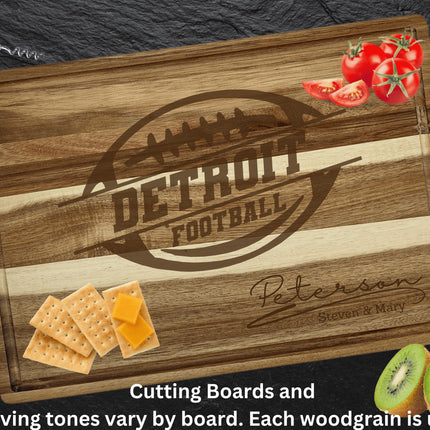 “Personalized Detroit football cutting board made from striped acacia wood, engraved with a football logo and the family name ‘Peterson – Steven & Mary.’ Displayed on a dark slate background with tomatoes, crackers with cheese, and sliced kiwi around it. Text at the bottom reads, ‘Cutting boards and engraving tones vary by board. Each woodgrain is unique.’”