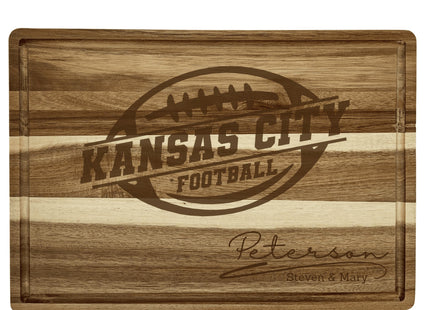 Alt text: Personalized acacia wood cutting board engraved with the words “Kansas City Football” inside a football logo and custom text below reading “Peterson – Steven & Mary.” The board features a natural striped woodgrain pattern with a deep juice groove around the edges.