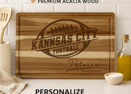 Custom acacia wood cutting board engraved with the words “Kansas City Football” inside a football logo, personalized below with the family name “Peterson” and “Steven & Mary.” The board is displayed upright on a white kitchen counter with a white tile backsplash, surrounded by kitchen utensils, a bottle of olive oil, and a small bowl of salt. Text above reads “CUSTOM CUTTING BOARD • PREMIUM ACACIA WOOD,” and below reads “PERSONALIZE WITH YOUR NAME.”