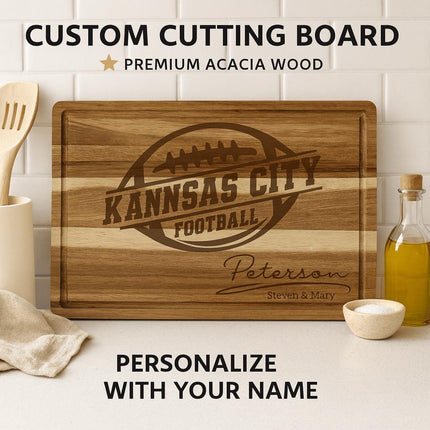 Custom acacia wood cutting board engraved with the words “Kansas City Football” inside a football logo, personalized below with the family name “Peterson” and “Steven & Mary.” The board is displayed upright on a white kitchen counter with a white tile backsplash, surrounded by kitchen utensils, a bottle of olive oil, and a small bowl of salt. Text above reads “CUSTOM CUTTING BOARD • PREMIUM ACACIA WOOD,” and below reads “PERSONALIZE WITH YOUR NAME.”