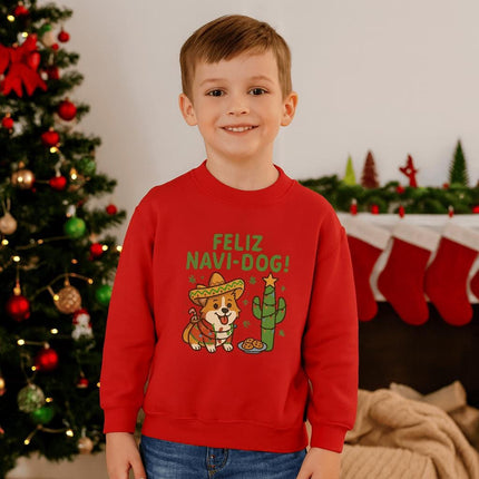 Smiling young boy standing in front of a decorated Christmas tree and fireplace, wearing a red sweatshirt with a festive “Feliz Navi-Dog!” design. The graphic features a cheerful Corgi wearing a sombrero and poncho next to a cactus decorated like a Christmas tree with a star on top and cookies nearby. Christmas stockings and holiday décor are visible in the background.