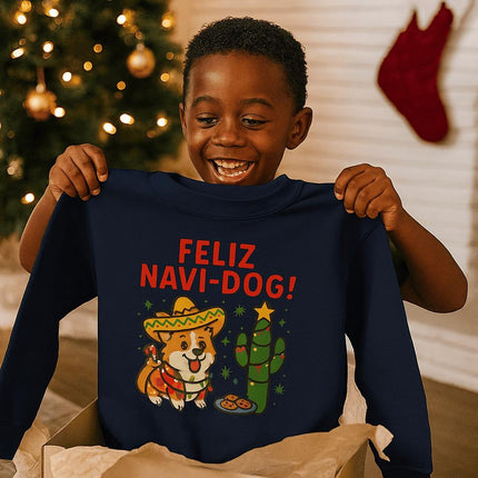 Excited young boy smiling as he holds up a navy blue sweatshirt with a festive “Feliz Navi-Dog!” design. The graphic features a cheerful Corgi wearing a sombrero and poncho next to a cactus decorated like a Christmas tree with a star on top and cookies nearby. A Christmas tree with gold ornaments and stockings hanging on the wall are visible in the cozy holiday background.