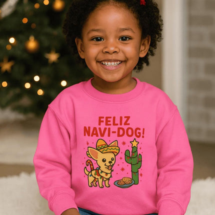 Smiling young girl sitting in front of a decorated Christmas tree wearing a bright pink sweatshirt with a festive “Feliz Navi-Dog!” design. The graphic features an adorable Chihuahua wearing a sombrero and scarf beside a cactus decorated like a Christmas tree with a star on top and a plate of cookies nearby. Warm holiday lights and ornaments create a cozy background.