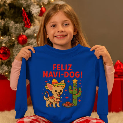 Smiling young girl sitting by a decorated Christmas tree holding up a royal blue sweatshirt with a festive “Feliz Navi-Dog!” design. The graphic features a cute Chihuahua wearing a sombrero and scarf beside a cactus decorated like a Christmas tree with a star on top and cookies nearby. Red ornaments and wrapped gifts are visible in the cozy holiday background.