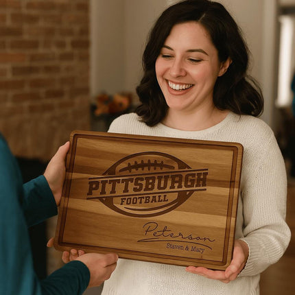 Smiling woman receives a personalized acacia wood cutting board engraved with “Pittsburgh Football” and “Peterson – Steven & Mary.” The board features a football design and engraved text, presented as a thoughtful gift in a cozy home setting with warm lighting and a brick wall in the background.