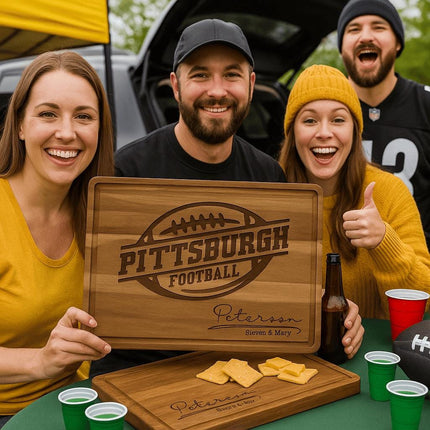 Smiling group of football fans at a Pittsburgh tailgate party holding a personalized acacia wood cutting board engraved with “Pittsburgh Football” and “Peterson – Steven & Mary.” The group is dressed in black and gold team colors, surrounded by red and green party cups, snacks, and a football, celebrating outdoors near parked vehicles.