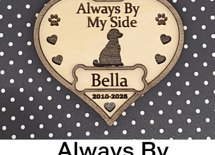 Heart-shaped wooden dog memorial ornament engraved with “Always By My Side,” featuring a dog silhouette, paw prints, and hearts. The ornament includes the name “Bella” and dates “2010–2025,” with a white ribbon for hanging, displayed against a black background with white polka dots. Text below reads “Always By My Side.”