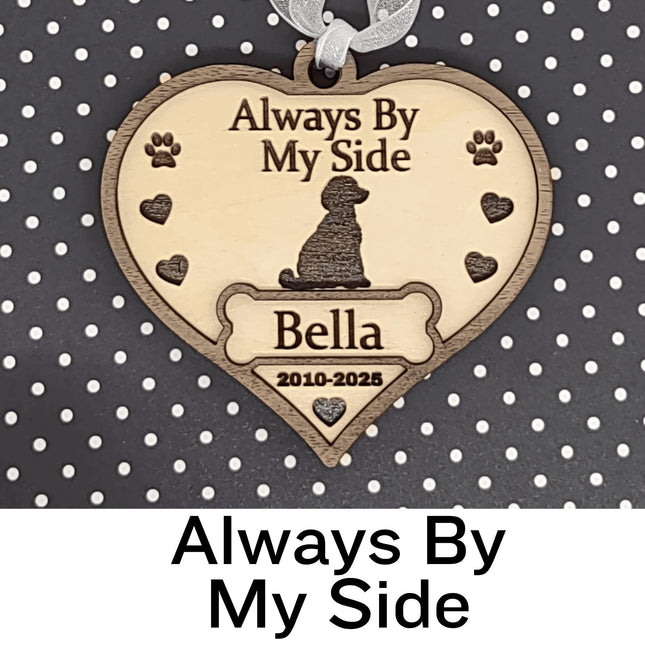 Heart-shaped wooden dog memorial ornament engraved with “Always By My Side,” featuring a dog silhouette, paw prints, and hearts. The ornament includes the name “Bella” and dates “2010–2025,” with a white ribbon for hanging, displayed against a black background with white polka dots. Text below reads “Always By My Side.”