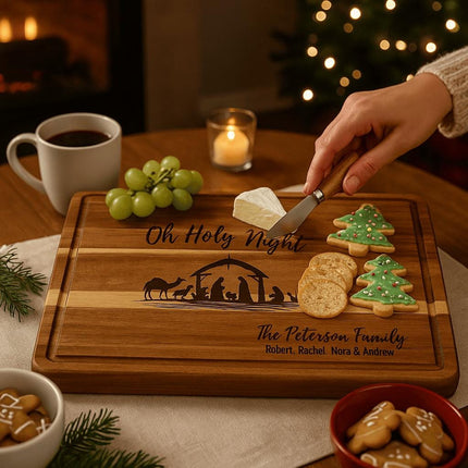 Personalized acacia cutting board engraved with a Nativity scene and the words “Oh Holy Night” and “The Peterson Family” is used to serve Christmas cookies, crackers, grapes, and cheese, with a hand spreading cheese and a holiday background featuring a candle, coffee, and Christmas tree lights.