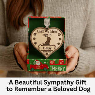 A woman holds an open festive gift box containing a personalized wooden dog memorial ornament engraved with “Until We Meet Again,” featuring a dog silhouette, paw prints, hearts, and the name “Daisy” with dates “2012–2028.” The box has a red and green Christmas design, and text below reads “A Beautiful Sympathy Gift to Remember a Beloved Dog.”