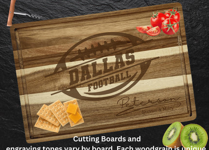 Alt text: “Dallas Football custom acacia cutting board engraved with a football logo and the name ‘Peterson – Steven & Mary.’ The board rests on a dark slate surface with tomatoes, crackers with cheese, and sliced kiwi arranged around it. Text at the bottom reads: ‘Cutting Boards and engraving tones vary by board. Each woodgrain is unique.’”