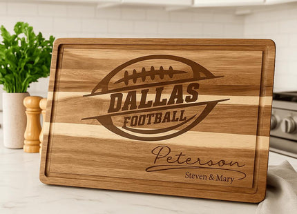 “Personalized Dallas Football acacia cutting board engraved with a football logo and the name ‘Peterson – Steven & Mary,’ displayed on a white marble kitchen countertop. The background features a bright modern kitchen with white cabinets, a green plant, wooden salt and pepper shakers, and a soft towel beside the board.”
