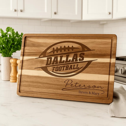 “Personalized Dallas Football acacia cutting board engraved with a football logo and the name ‘Peterson – Steven & Mary,’ displayed on a white marble kitchen countertop. The background features a bright modern kitchen with white cabinets, a green plant, wooden salt and pepper shakers, and a soft towel beside the board.”