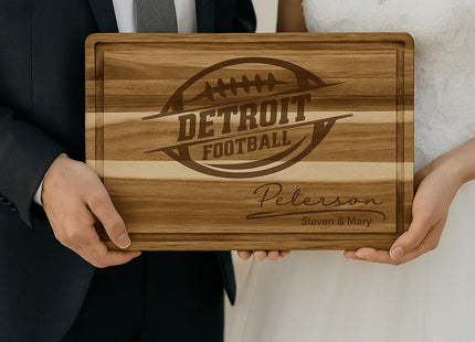 “Bride and groom holding a personalized Detroit football cutting board engraved with a football logo and the family name ‘Peterson – Steven & Mary.’ The acacia wood board features a striped grain pattern and juice groove, making it a unique and thoughtful wedding gift for football fans.”