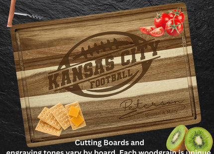 Personalized acacia wood cutting board engraved with “Kansas City Football” inside a football logo and “Peterson – Steven & Mary” below. The board rests on a dark slate surface with crackers and cheese in the lower left corner, sliced kiwis in the lower right, tomatoes in the top right, and a corkscrew at the top left. Text at the bottom reads, “Cutting Boards and engraving tones vary by board. Each woodgrain is unique.”