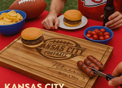 Alt text: Personalized acacia wood cutting board engraved with “Kansas City Football” and the name “Peterson – Steven & Mary,” displayed on a red tablecloth during a Kansas City football game day cookout. The scene includes burgers, grilled sausages, a bowl of chips, cherry tomatoes, a football, and fans wearing red Kansas City shirts. Text in the bottom left corner reads “KANSAS CITY GAME DAY.”