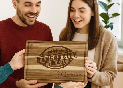 A smiling couple receives a personalized acacia wood cutting board engraved with “Kansas City Football” and the name “Peterson – Steven & Mary.” The man wears a red sweater, and the woman wears a beige cardigan. They appear delighted as another person hands them the board in a bright, cozy indoor setting with moving boxes and a plant in the background.