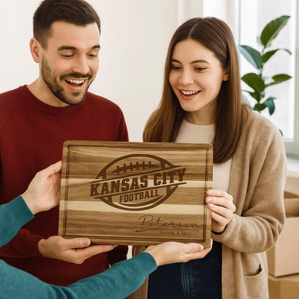 A smiling couple receives a personalized acacia wood cutting board engraved with “Kansas City Football” and the name “Peterson – Steven & Mary.” The man wears a red sweater, and the woman wears a beige cardigan. They appear delighted as another person hands them the board in a bright, cozy indoor setting with moving boxes and a plant in the background.
