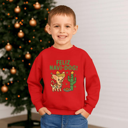Smiling young boy standing in front of a decorated Christmas tree wearing a red sweatshirt with a festive “Feliz Navi-Dog!” design. The graphic shows a cheerful Chihuahua wearing a sombrero and scarf beside a cactus decorated like a Christmas tree with a star on top and cookies nearby. Warm holiday lights and gold ornaments create a cozy Christmas background.