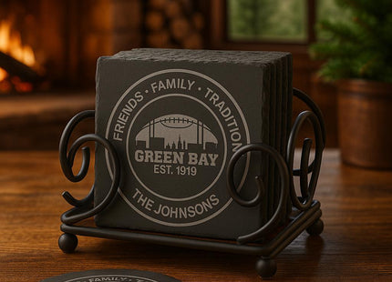 &quot;Set of personalized Green Bay slate coasters displayed in a black iron holder on a wooden table. The engraving reads &#39;Friends • Family • Tradition • Green Bay Est. 1919 • The Johnsons.&#39; A cozy cabin scene with a fireplace and pine trees is blurred in the background, creating a warm, rustic atmosphere.&quot;