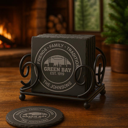 "Set of personalized Green Bay slate coasters displayed in a black iron holder on a wooden table. The engraving reads 'Friends • Family • Tradition • Green Bay Est. 1919 • The Johnsons.' A cozy cabin scene with a fireplace and pine trees is blurred in the background, creating a warm, rustic atmosphere."