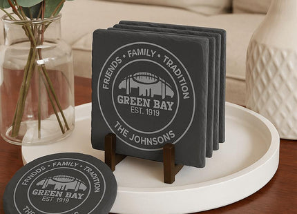 &quot;Set of personalized Green Bay slate coasters displayed on a modern coffee table. The engraving reads &#39;Friends • Family • Tradition • Green Bay Est. 1919 • The Johnsons.&#39; Coasters are stacked neatly in a dark walnut wood holder on a white round tray, with one coaster placed in front. A glass vase with eucalyptus stems and a cream-colored sofa are in the background, creating a cozy home setting.&quot;