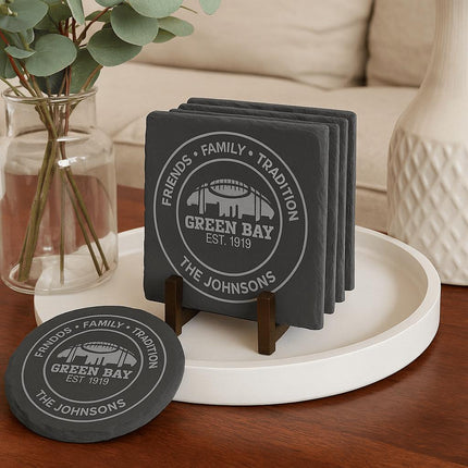 "Set of personalized Green Bay slate coasters displayed on a modern coffee table. The engraving reads 'Friends • Family • Tradition • Green Bay Est. 1919 • The Johnsons.' Coasters are stacked neatly in a dark walnut wood holder on a white round tray, with one coaster placed in front. A glass vase with eucalyptus stems and a cream-colored sofa are in the background, creating a cozy home setting."