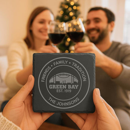 "Close-up of a personalized Green Bay slate coaster engraved with 'Friends • Family • Tradition,' 'Green Bay Est. 1919,' and 'The Johnsons.' In the background, a smiling couple clinks wine glasses near a decorated Christmas tree, creating a cozy holiday atmosphere."