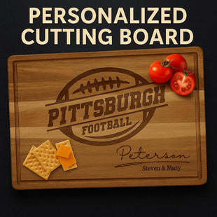 Personalized acacia wood cutting board engraved with a football design and the text “Pittsburgh Football.” Below, it features the family name “Peterson” in elegant script with “Steven & Mary” engraved underneath. The board has a juice groove around the edge and is styled with sliced tomatoes and cheese crackers for a warm, rustic presentation.