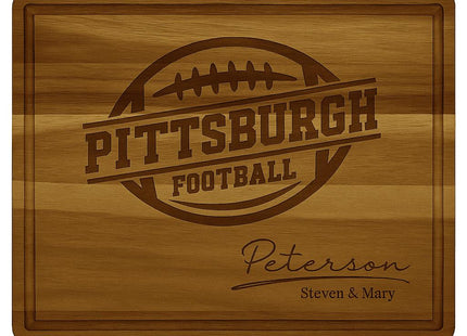 Personalized acacia wood cutting board engraved with the text “Pittsburgh Football” and a football graphic in the center. The bottom right corner features the family name “Peterson” in elegant script with “Steven & Mary” engraved below. The board has a smooth finish with a beveled juice groove around the edge, highlighting its craftsmanship and rich wood grain.