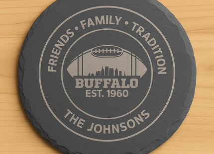 &quot;Round black slate coaster engraved with a Buffalo football design. The engraving features the text &#39;Friends • Family • Tradition,&#39; &#39;Buffalo Est. 1960,&#39; and &#39;The Johnsons,&#39; along with a football silhouette containing a city skyline. The coaster is displayed on a light wooden surface.&quot;