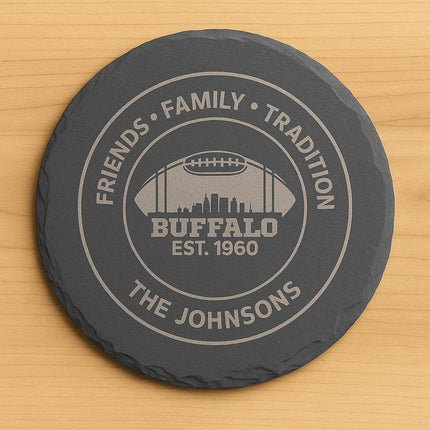 "Round black slate coaster engraved with a Buffalo football design. The engraving features the text 'Friends • Family • Tradition,' 'Buffalo Est. 1960,' and 'The Johnsons,' along with a football silhouette containing a city skyline. The coaster is displayed on a light wooden surface."