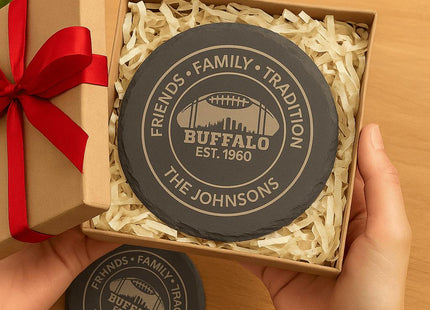 &quot;Hands holding an open gift box with personalized round black slate coasters engraved with &#39;Friends • Family • Tradition,&#39; &#39;Buffalo Est. 1960,&#39; and &#39;The Johnsons.&#39; The coasters are nestled in shredded kraft paper inside a brown gift box with a red ribbon bow, creating a festive presentation.&quot;