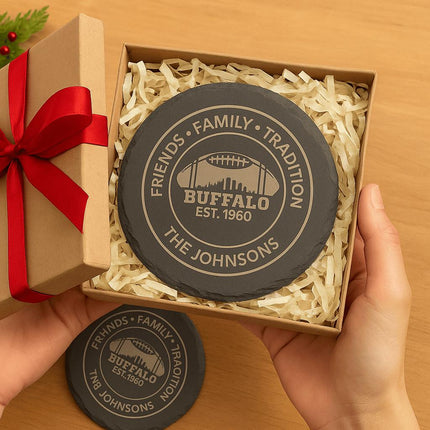 "Hands holding an open gift box with personalized round black slate coasters engraved with 'Friends • Family • Tradition,' 'Buffalo Est. 1960,' and 'The Johnsons.' The coasters are nestled in shredded kraft paper inside a brown gift box with a red ribbon bow, creating a festive presentation."