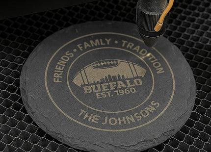 &quot;Round black slate coaster being laser engraved with the text &#39;Friends • Family • Tradition,&#39; &#39;Buffalo Est. 1960,&#39; and &#39;The Johnsons,&#39; featuring a football and city skyline design. The coaster sits on a honeycomb laser bed under the laser engraving machine in action.&quot;