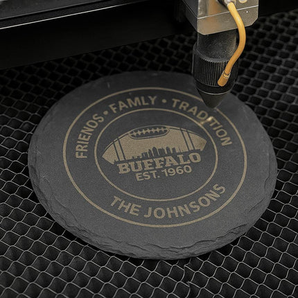 "Round black slate coaster being laser engraved with the text 'Friends • Family • Tradition,' 'Buffalo Est. 1960,' and 'The Johnsons,' featuring a football and city skyline design. The coaster sits on a honeycomb laser bed under the laser engraving machine in action."