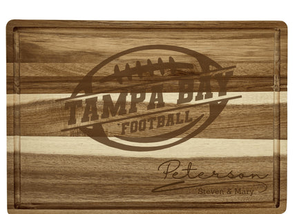 Personalized acacia wood cutting board engraved with a football logo and the text “Tampa Bay Football.” The bottom right corner features the engraved family name “Peterson” with “Steven & Mary” below it. The board shows natural striped wood grain and a carved juice groove around the edge.