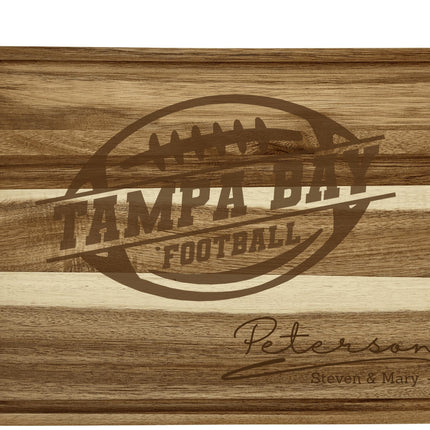Personalized acacia wood cutting board engraved with a football logo and the text “Tampa Bay Football.” The bottom right corner features the engraved family name “Peterson” with “Steven & Mary” below it. The board shows natural striped wood grain and a carved juice groove around the edge.
