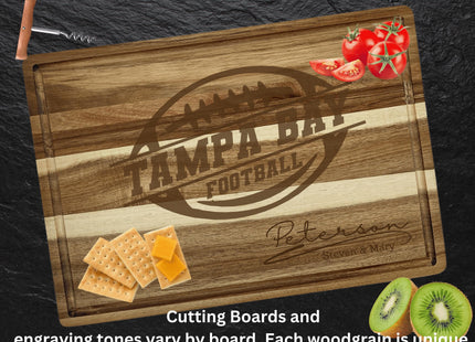 Personalized acacia wood cutting board engraved with a football logo and the text “Tampa Bay Football,” along with the family name “Peterson – Steven & Mary.” The board is styled on a dark stone surface with tomatoes in the top right corner, crackers and cheese in the bottom left, and sliced kiwi in the bottom right. A corkscrew is placed at the top left. Text at the bottom reads, “Cutting boards and engraving tones vary by board. Each woodgrain is unique.”