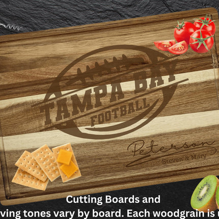 Personalized acacia wood cutting board engraved with a football logo and the text “Tampa Bay Football,” along with the family name “Peterson – Steven & Mary.” The board is styled on a dark stone surface with tomatoes in the top right corner, crackers and cheese in the bottom left, and sliced kiwi in the bottom right. A corkscrew is placed at the top left. Text at the bottom reads, “Cutting boards and engraving tones vary by board. Each woodgrain is unique.”