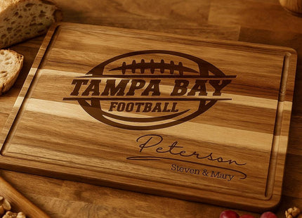 Personalized acacia wood cutting board engraved with “Tampa Bay Football” and the family name “Peterson – Steven & Mary,” displayed on a kitchen countertop. The board is surrounded by fresh bread, walnuts, cheese, grapes, and rosemary, highlighting its use as a serving or charcuterie board.