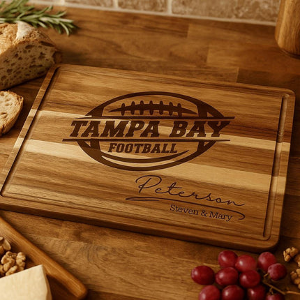 Personalized acacia wood cutting board engraved with “Tampa Bay Football” and the family name “Peterson – Steven & Mary,” displayed on a kitchen countertop. The board is surrounded by fresh bread, walnuts, cheese, grapes, and rosemary, highlighting its use as a serving or charcuterie board.