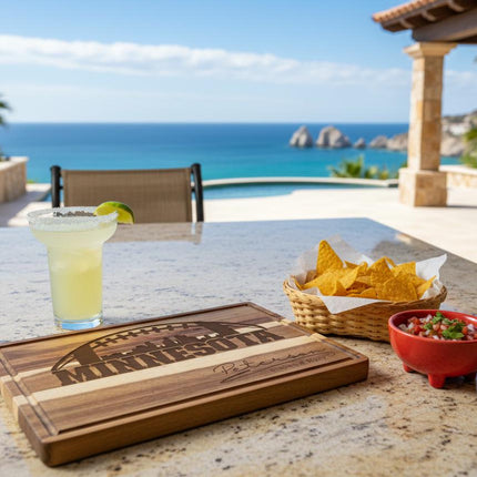 "Personalized Minnesota football cutting board engraved with a football and skyline design sits on an outdoor stone table overlooking a scenic ocean view. Next to it are a basket of tortilla chips, a red bowl of salsa, and a margarita with a lime wedge, creating a relaxed coastal entertaining scene."