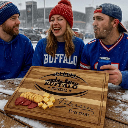 "Three Buffalo football fans dressed in team colors enjoying a snowy tailgate outside the stadium. In front of them on a picnic table is a personalized acacia cutting board engraved with a football design and 'Buffalo Football' logo, customized with the name 'Peterson.' The board displays sliced sausage, cheese cubes, and crackers, highlighting its use as a game-day serving board."
