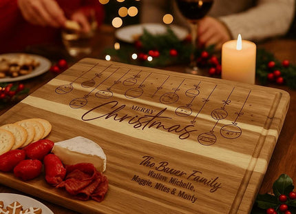 Personalized acacia cutting board engraved with hanging Christmas ornaments and the words “Merry Christmas” and “The Bauer Family” is displayed as a holiday charcuterie board with crackers, cheese, and meats, surrounded by candles, holly decor, and people enjoying food and wine in a festive setting.