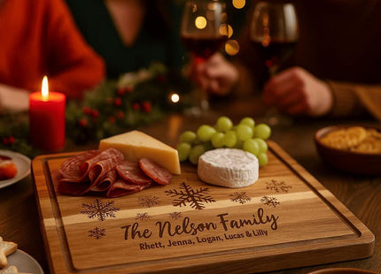 Personalized acacia cutting board engraved with snowflakes and “The Nelson Family” is used as a holiday charcuterie board holding cheese, salami, and grapes, with three people enjoying wine and Christmas lights in the background.