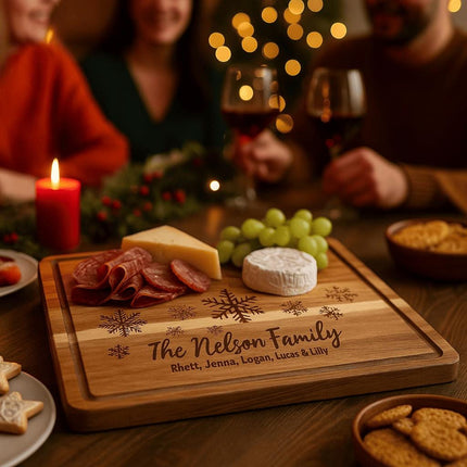 Personalized acacia cutting board engraved with snowflakes and “The Nelson Family” is used as a holiday charcuterie board holding cheese, salami, and grapes, with three people enjoying wine and Christmas lights in the background.