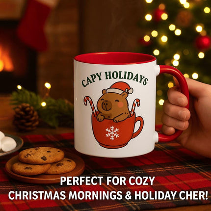 "Festive red-handled mug with a 'Capy Holidays' design featuring a smiling capybara in a red cup holding candy canes, surrounded by cozy holiday décor. The mug sits on a plaid tablecloth beside a plate of chocolate chip cookies and marshmallows, with a glowing Christmas tree and fireplace in the background. Text reads 'Perfect for Cozy Christmas Mornings & Holiday Cheer!'"